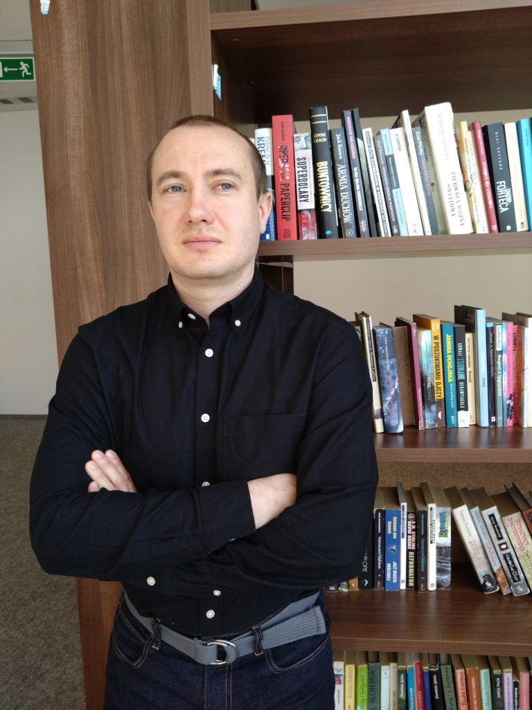 Zdjęcie dr Dariusza Kardeli na tle regału z książkami.